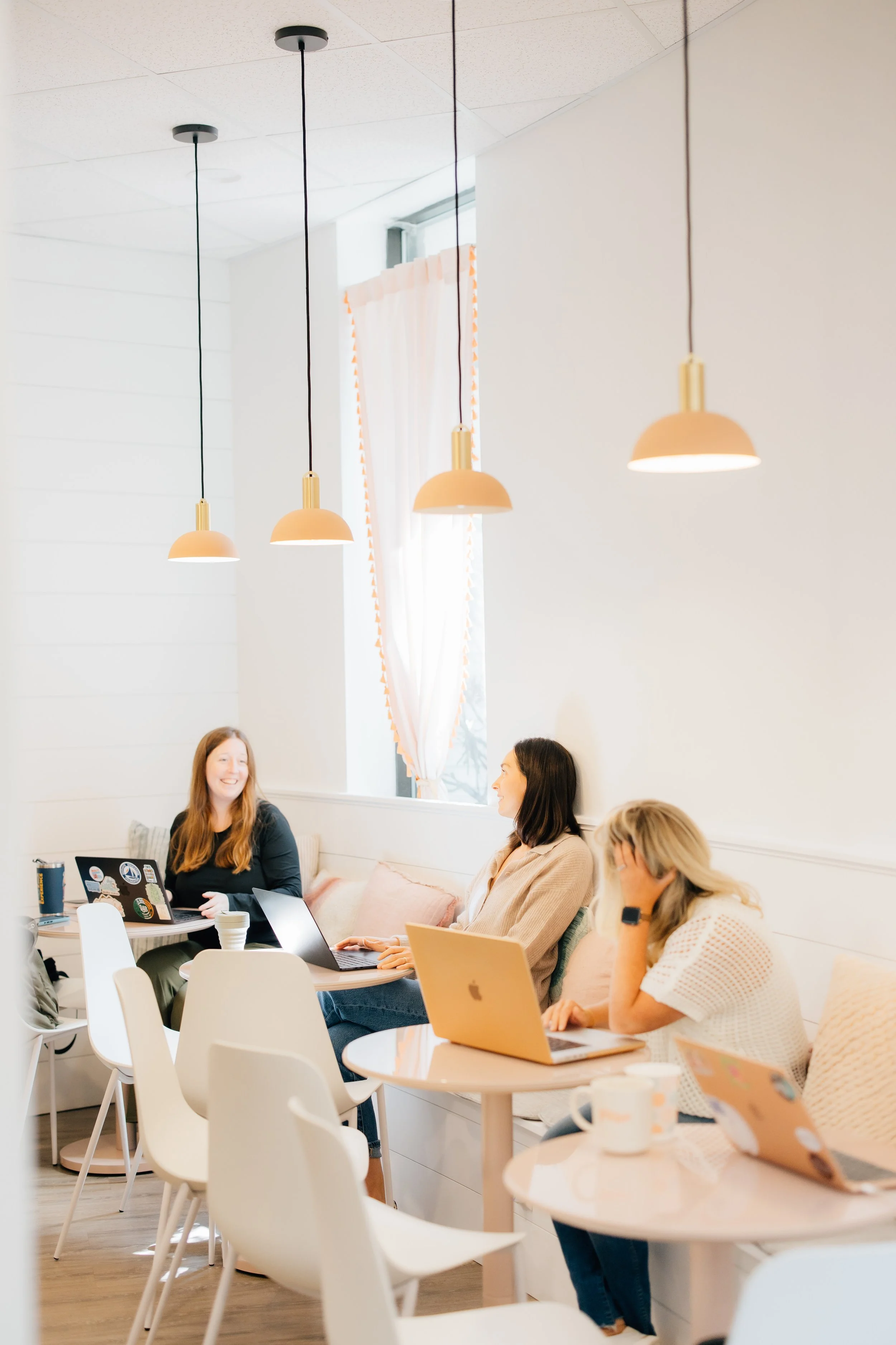 A group of women sitting at a round table in a brightly lit room with hanging pendant lights, working on laptops and having a discussion.