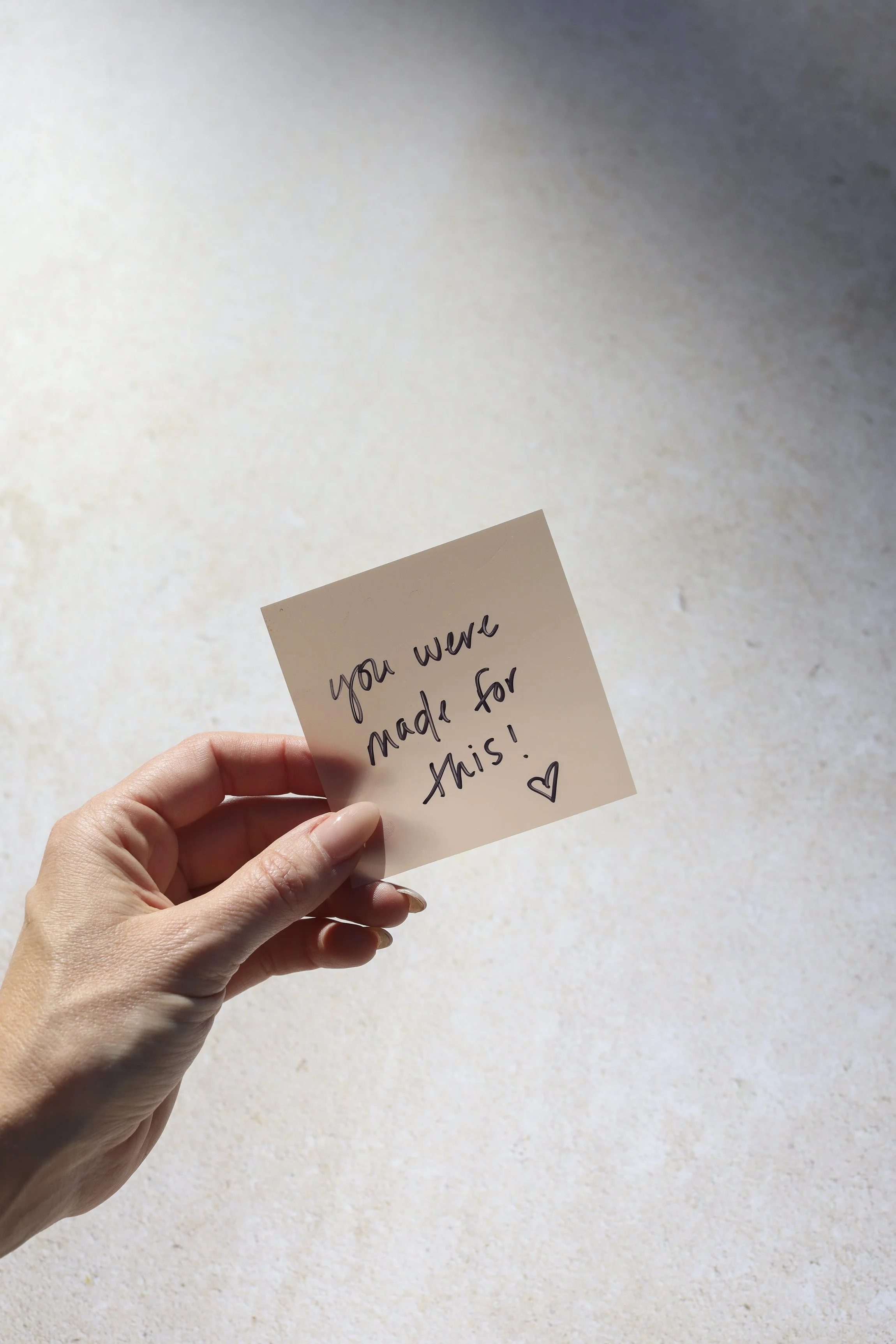 A person holding a small note card with handwritten message that reads 'You were made for this!' with a small heart at the bottom right corner. The background is a plain, light-colored surface.