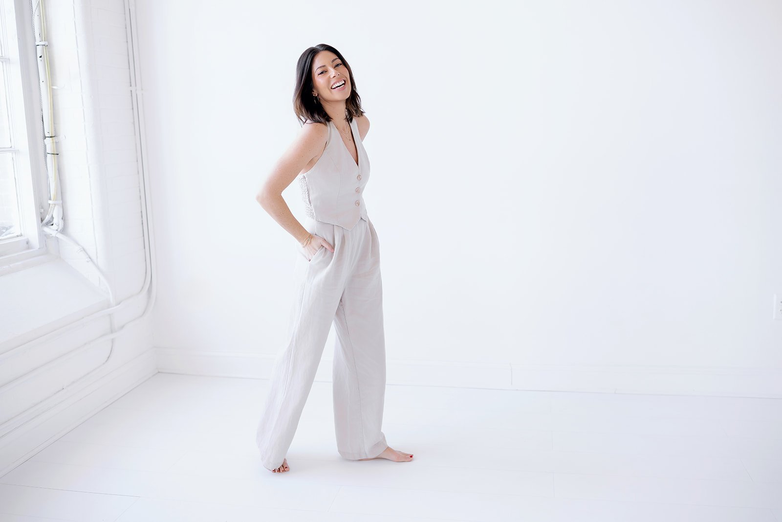 Woman wearing a sleeveless beige outfit, standing in a bright white room with natural light, smiling with one hand in her pocket.