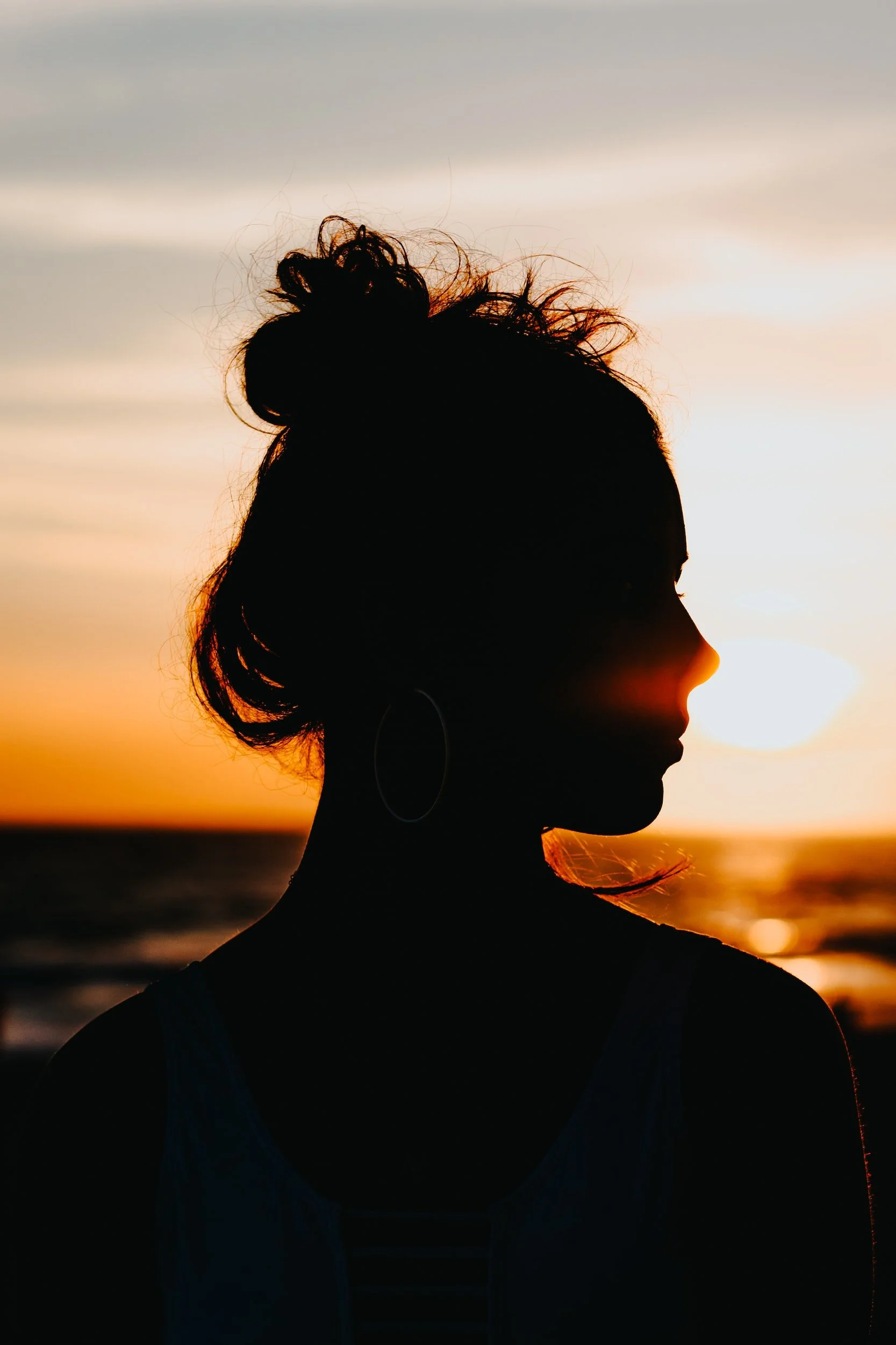 Silhouette of a woman with a bun hairstyle wearing a tank top and hoop earrings, with a sunset and ocean in the background.