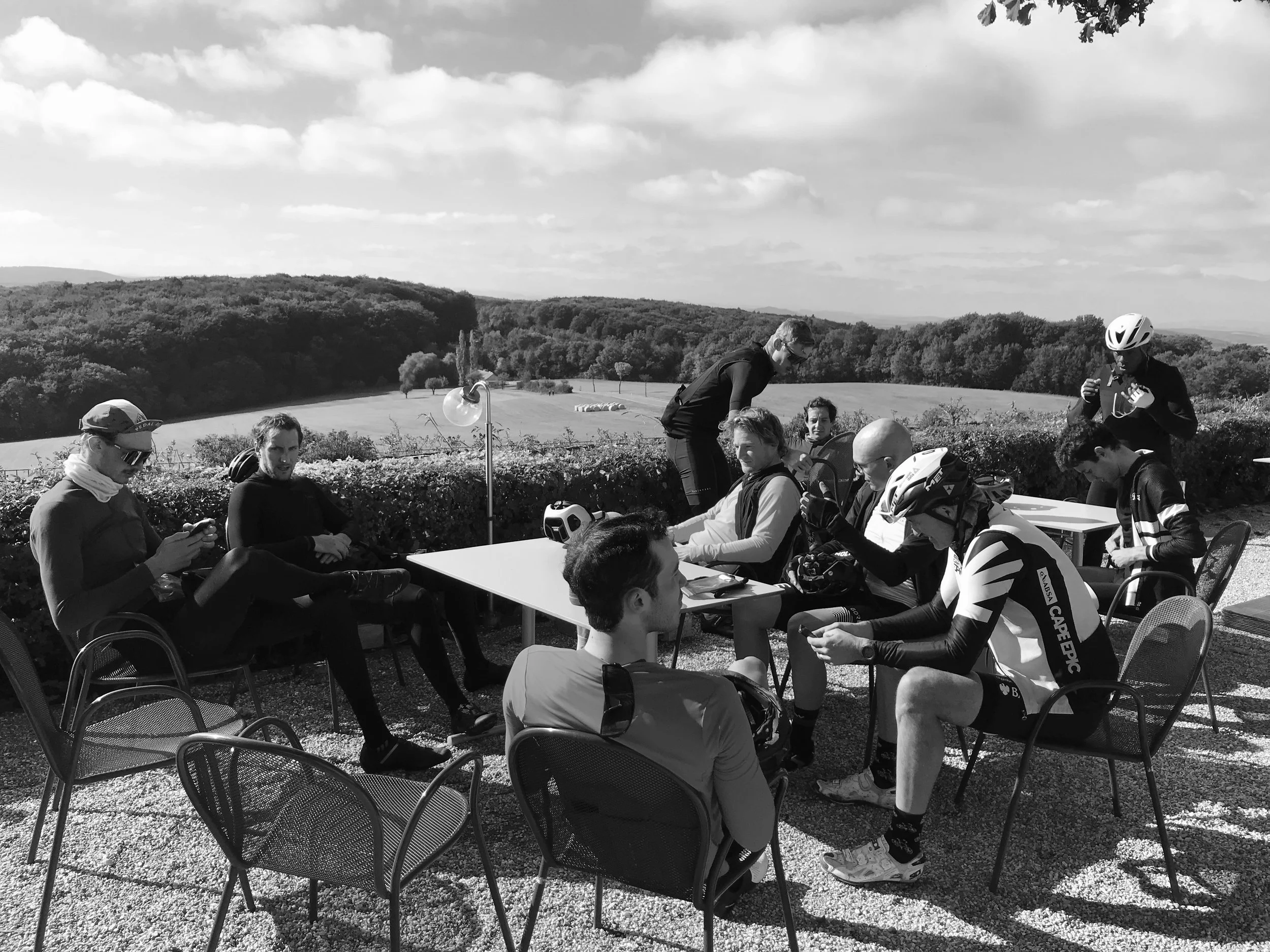 Group of cyclists sitting and standing outdoors at tables, some wearing helmets, with open countryside and rolling hills in the background.