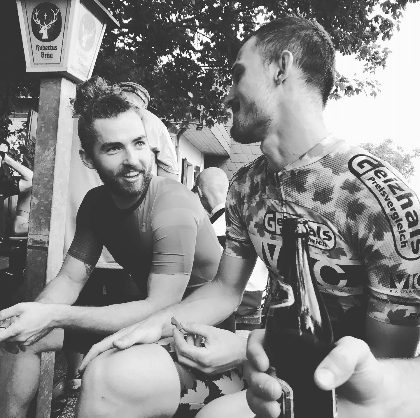 Two young men sitting outdoors, engaged in conversation. The man on the left has a beard and is smiling, wearing a cycling jersey. The man on the right has short hair, a beard, and is holding a beer bottle, also dressed in a cycling jersey. There are other people and trees in the background.