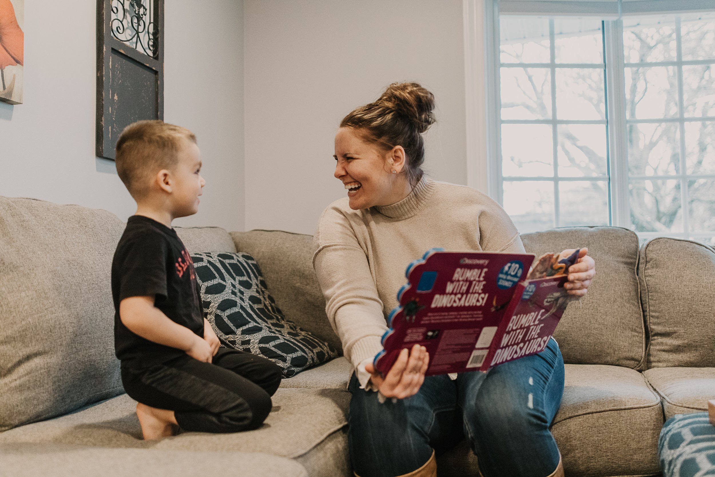 Caitlin Bergman smiles while reading an interactive dinosaur book with a young boy on a comfortable sofa.