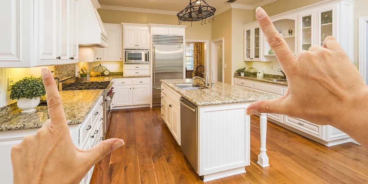 Hands framing a modern kitchen renovation with white cabinets, granite countertops, stainless steel appliances, and warm hardwood floors.