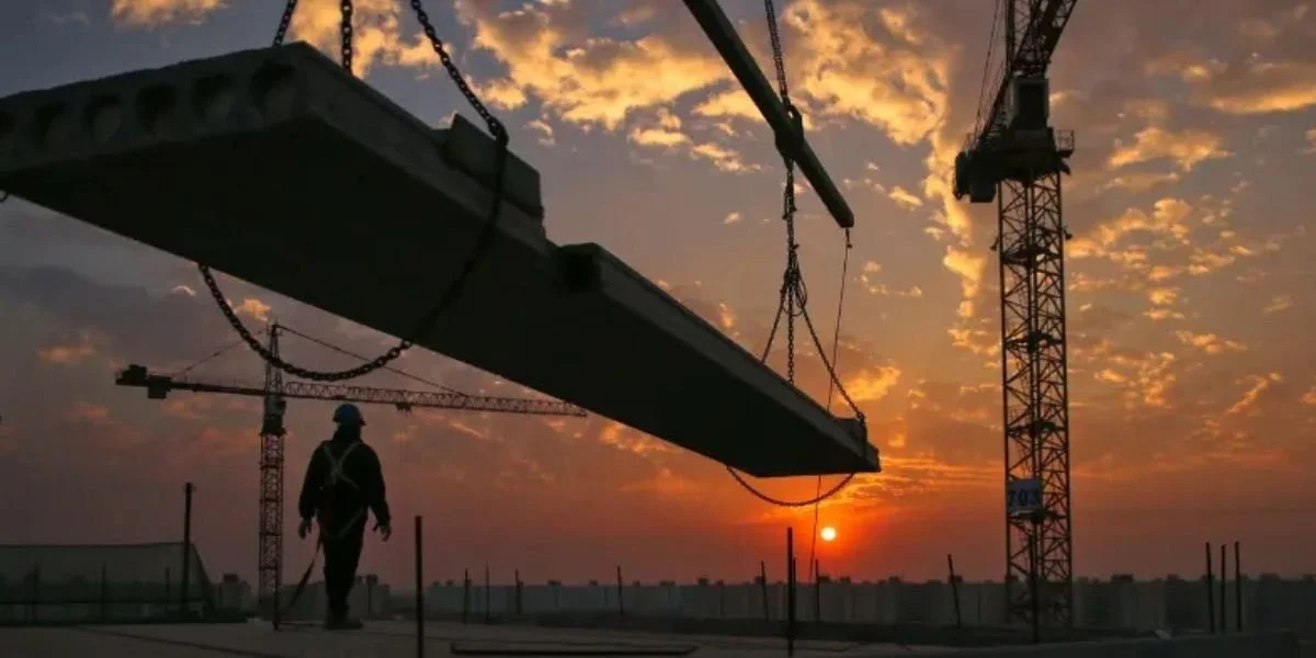 Construction site at sunset with cranes lifting heavy materials and a worker overseeing operations.