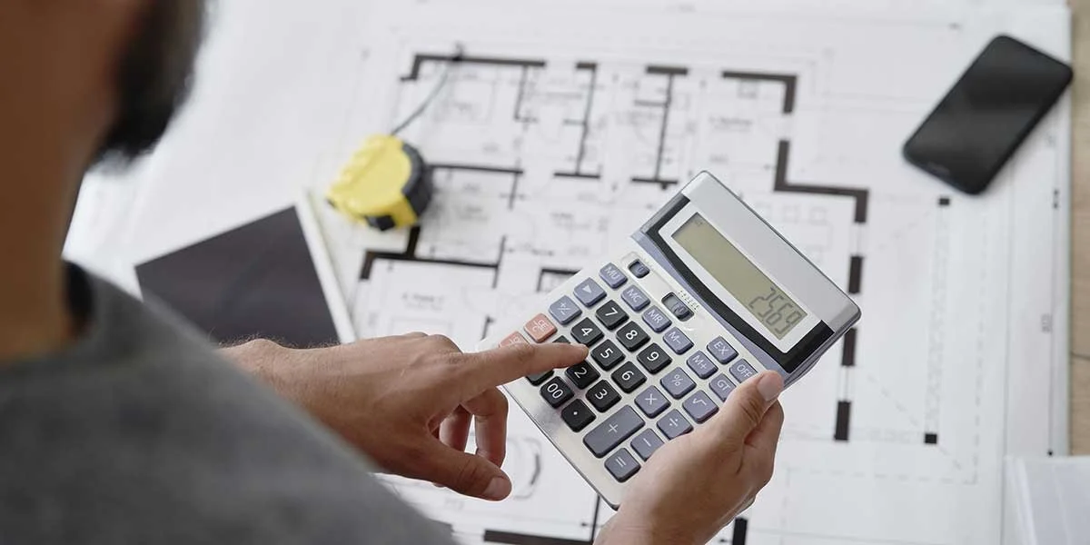 Person using a calculator over house blueprints to plan and budget, avoiding cost overruns during home remodeling projects.