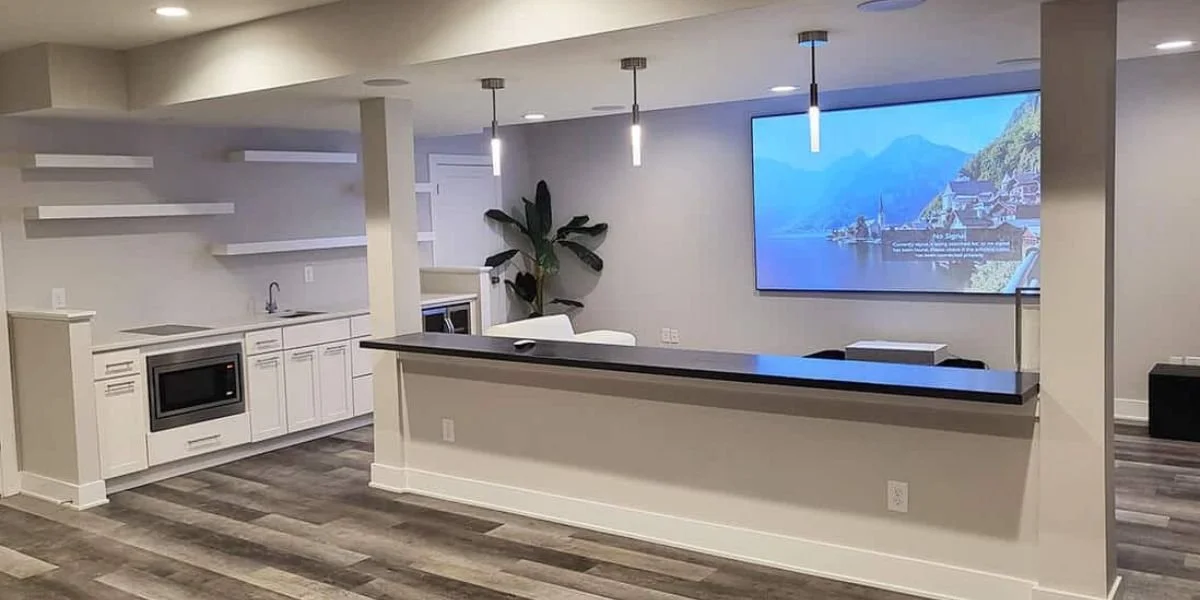 Basement remodeling project featuring a custom wet bar, white cabinetry, floating shelves, pendant lighting, and a large projection screen.
