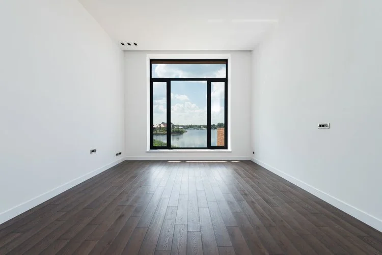 Dark wood-look laminate flooring extending across a minimalist room with white walls, white baseboards, and a black-framed window.