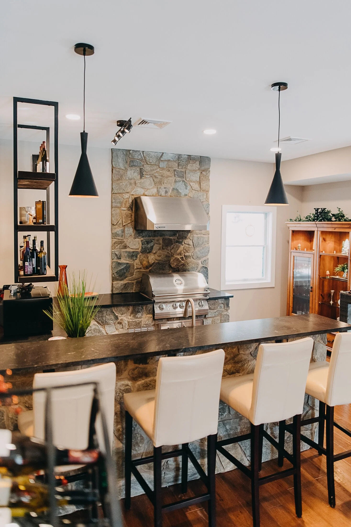 A luxury indoor kitchen bar features a dark stone countertop, stone accent wall, cream leather bar stools, and modern pendant lights.
