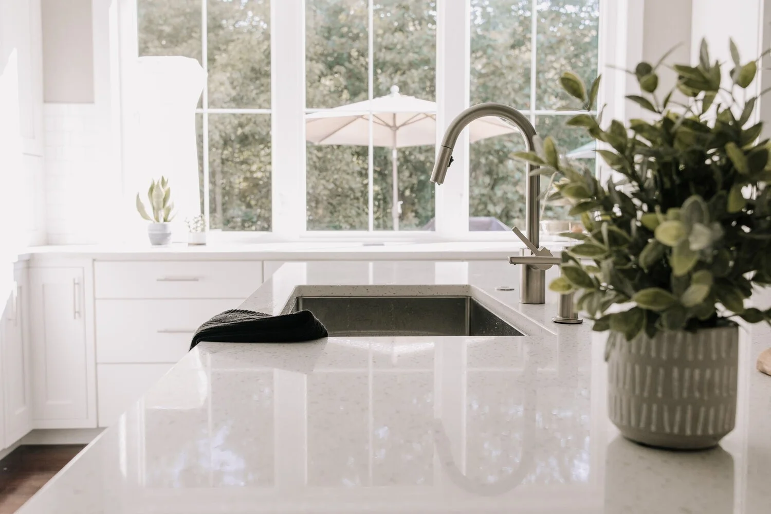 Elegant kitchen with understated white finishes, a polished quartz countertop, natural greenery, and soft daylight through a large window.