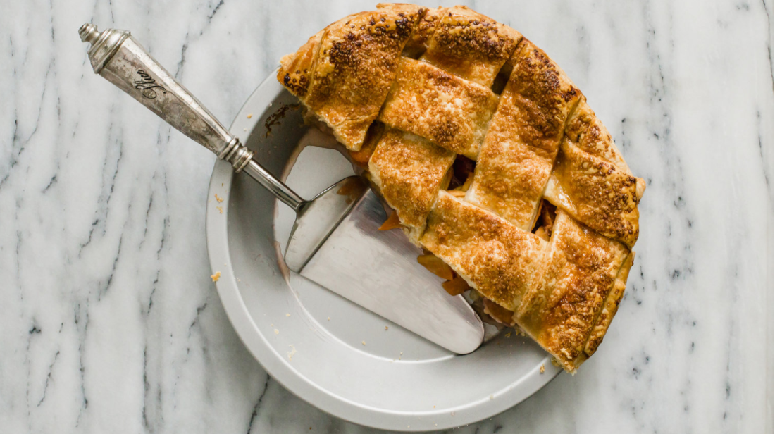 Freshly baked fruit pie with a golden-brown lattice crust sits in a pie pan on a white marble countertop.