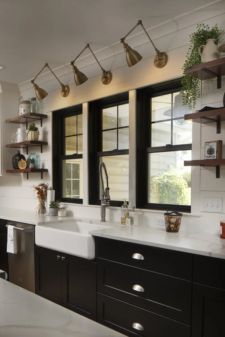 A modern farmhouse kitchen features dark lower cabinets, white quartz countertops, and a large white apron-front sink beneath three black-framed windows.