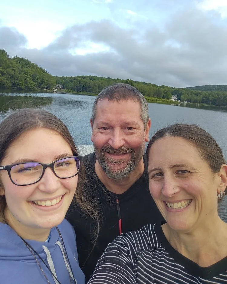 Kevin Kelly smiles with his wife and daughter in an outdoor selfie overlooking a scenic lake and forested hills.