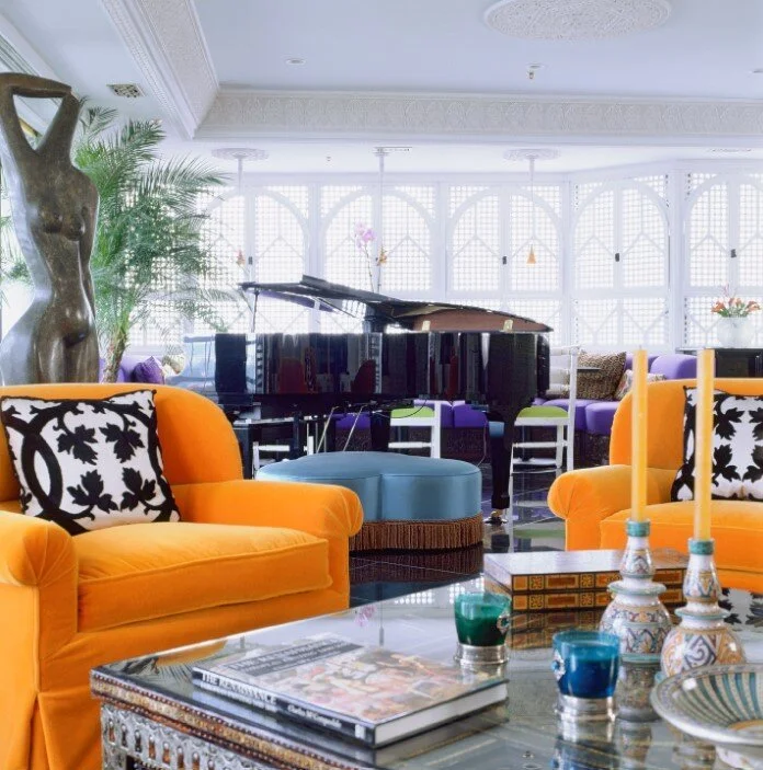 Luxury living room featuring bright orange velvet armchairs, a grand piano, a blue ottoman, and decorative coffee table accessories
