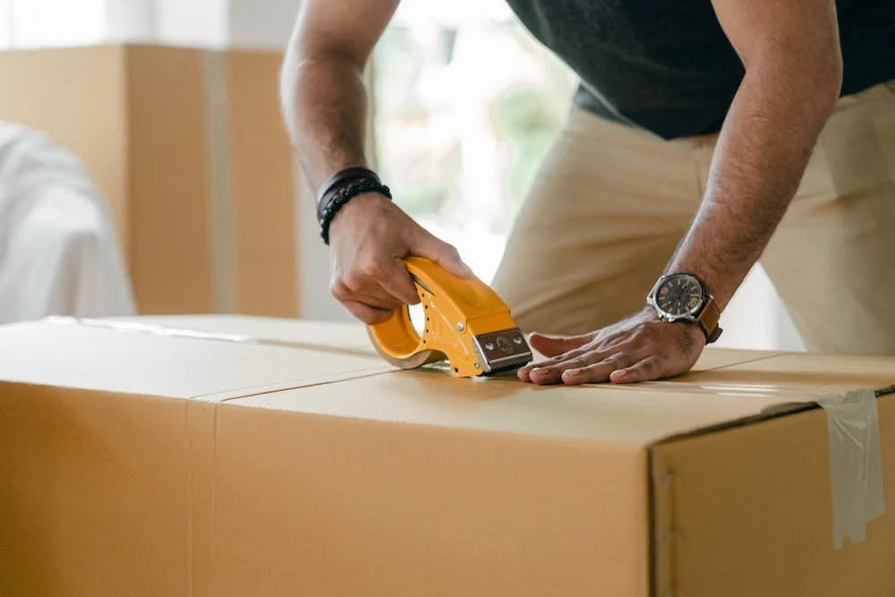 Close-up of a person taping a packed cardboard box with other shipping boxes visible in the background.