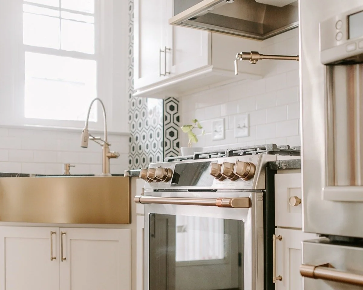 Modern white and bronze kitchen with a stainless steel gas range, matching hood, pot filler, and bronze-accented farmhouse sink.