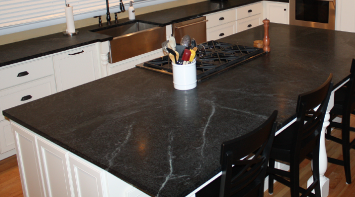 Large kitchen island featuring a dark Rocky Mountain Stone countertop with subtle light veining, a built-in cooktop, and black chairs.