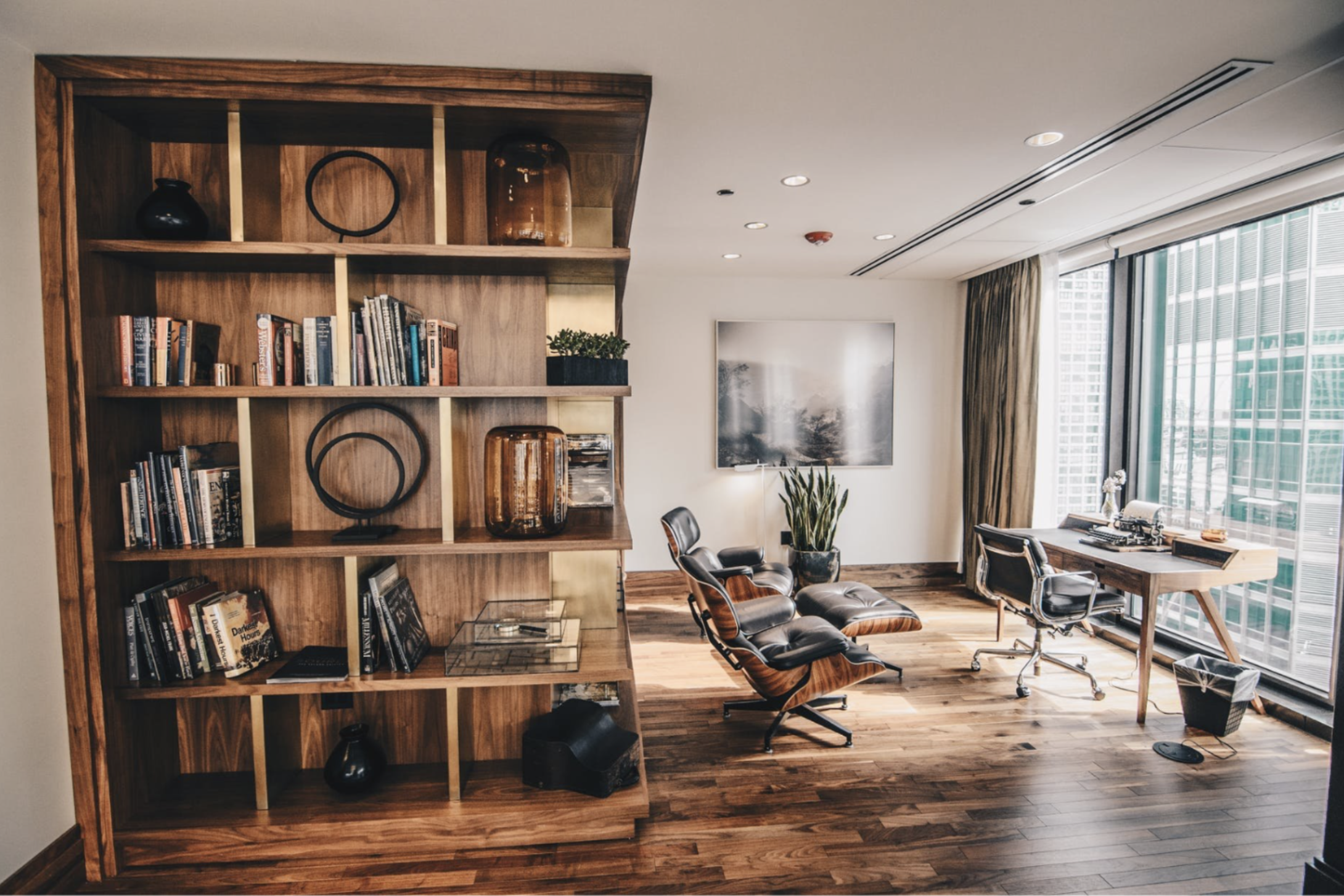 Spacious dedicated home office with a wooden desk, leather lounge chair, custom built-in bookcase, and large windows.