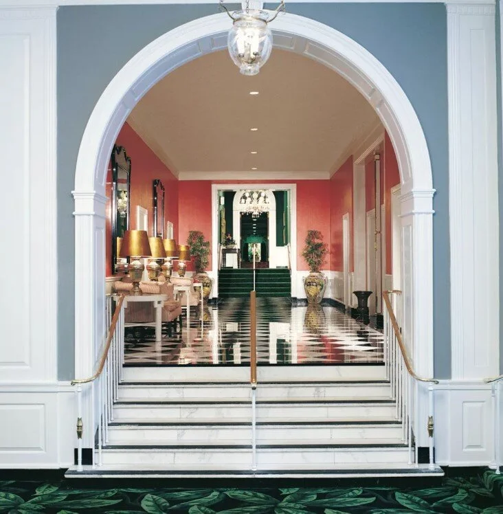 Elegant mansion interior featuring a grand marble staircase, checkered flooring, coral-red walls, and an arched entryway.