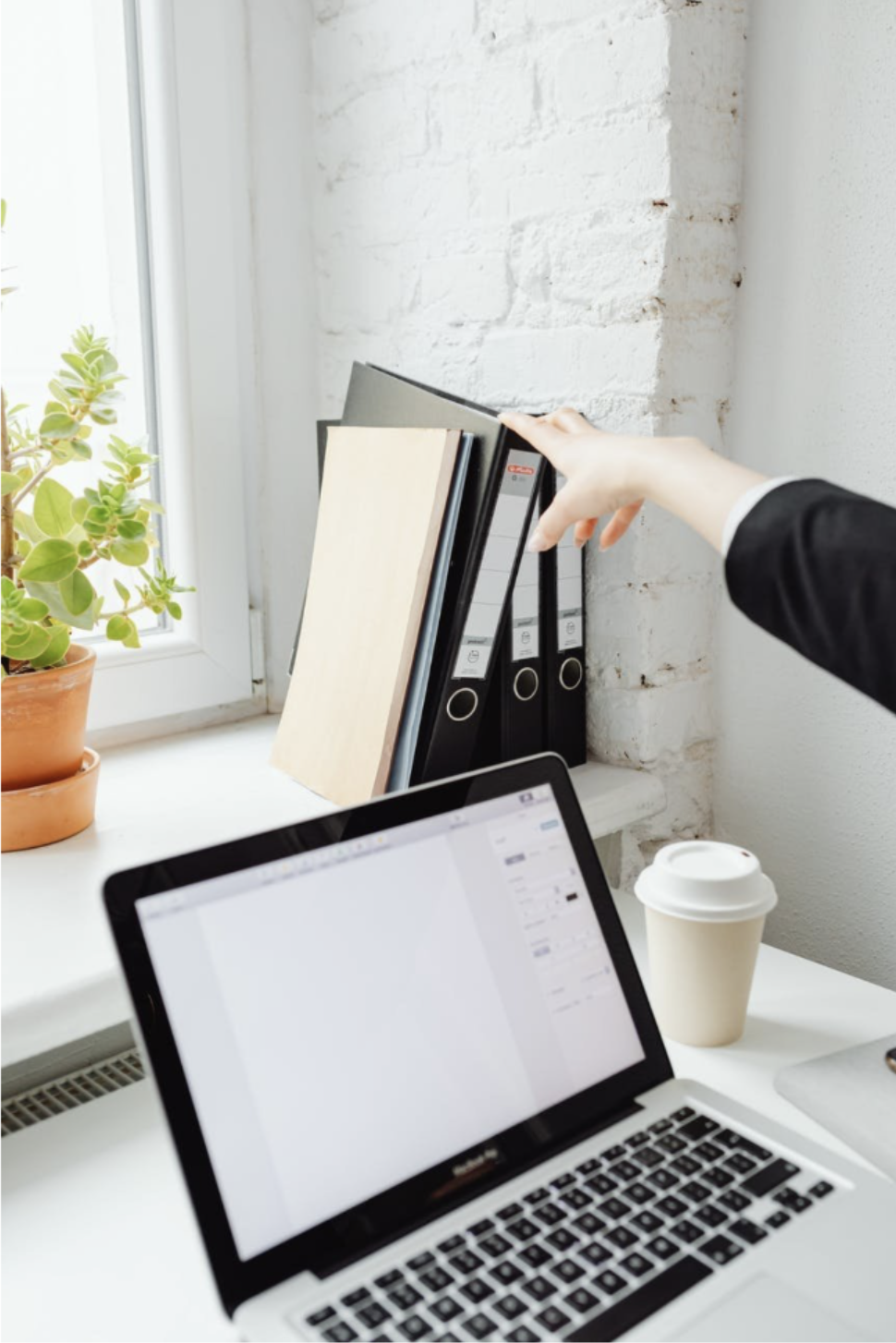 A hand selecting a binder from organized files beside a white desk with an open laptop and natural light.