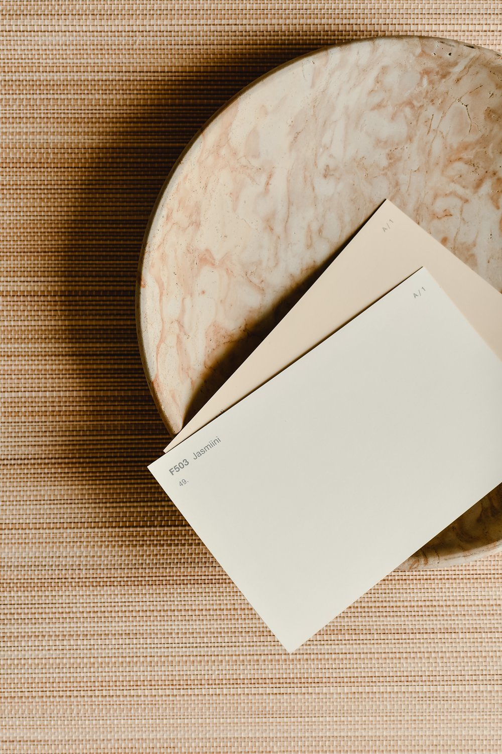 Neutral paint swatches in soft cream and beige tones sit in a marble bowl beside a textured woven placemat.