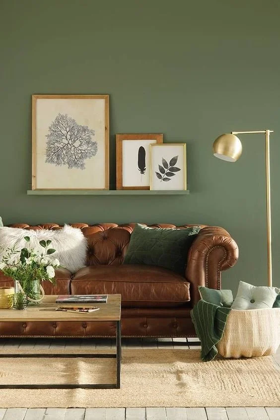 Cozy living room with an olive green accent wall, brown tufted leather sofa, jute rug, wooden coffee table, and botanical artwork.