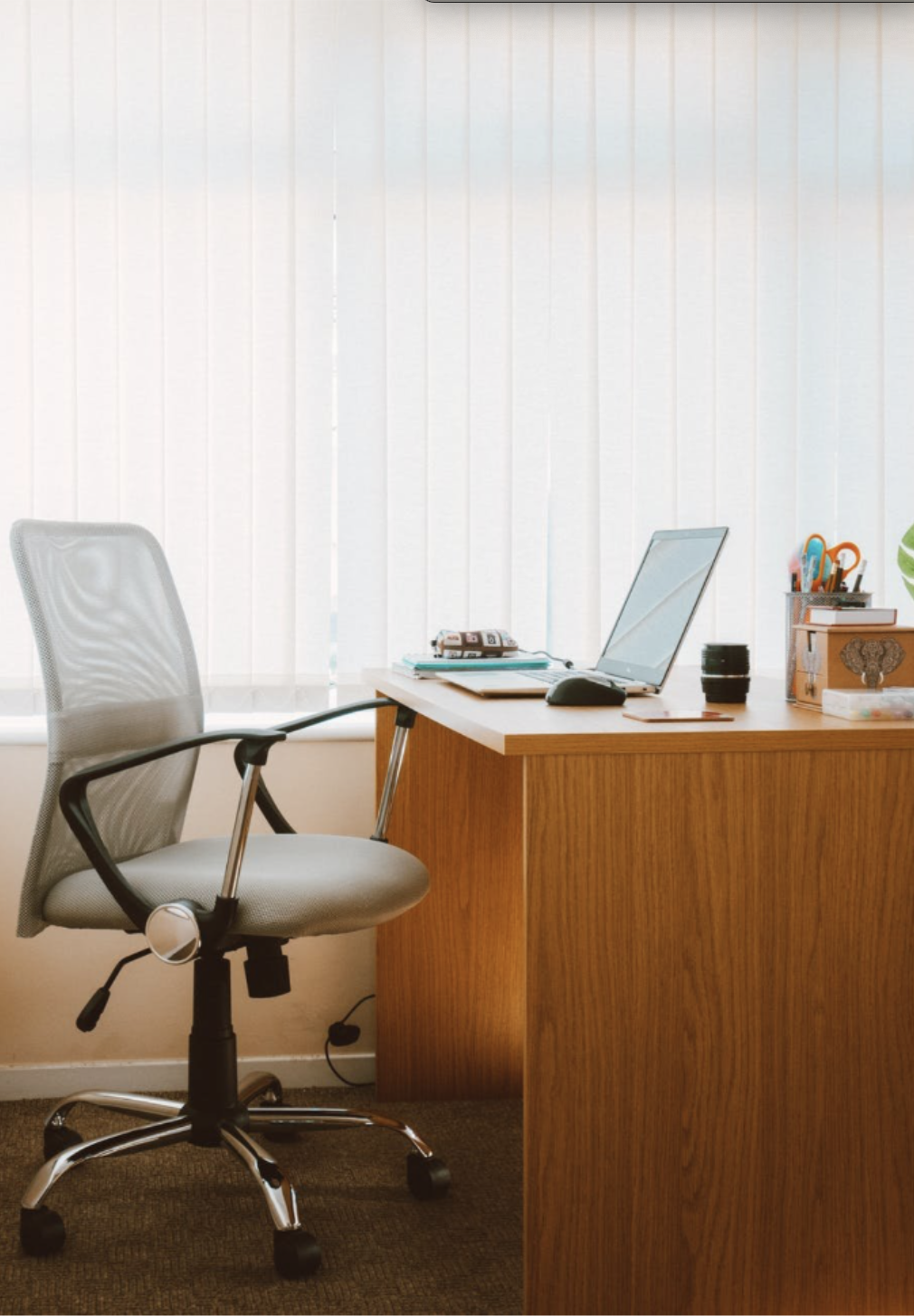 Home office with a wooden desk, open laptop, ergonomic gray mesh chair, and wall-to-wall carpeting for a quieter workspace.