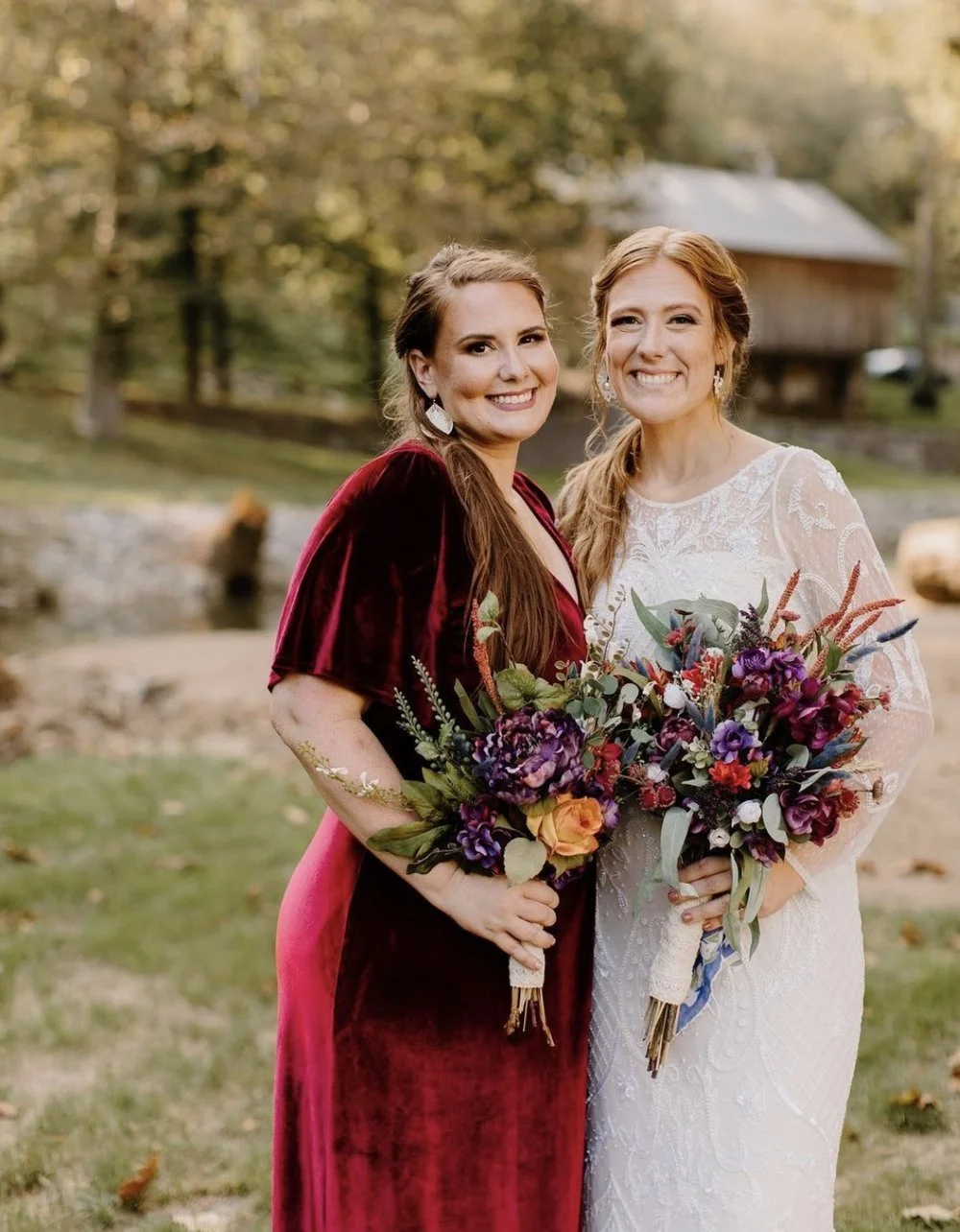 A bride in a white lace wedding dress and a bridesmaid in a deep red velvet dress pose together with colorful autumn bouquets.