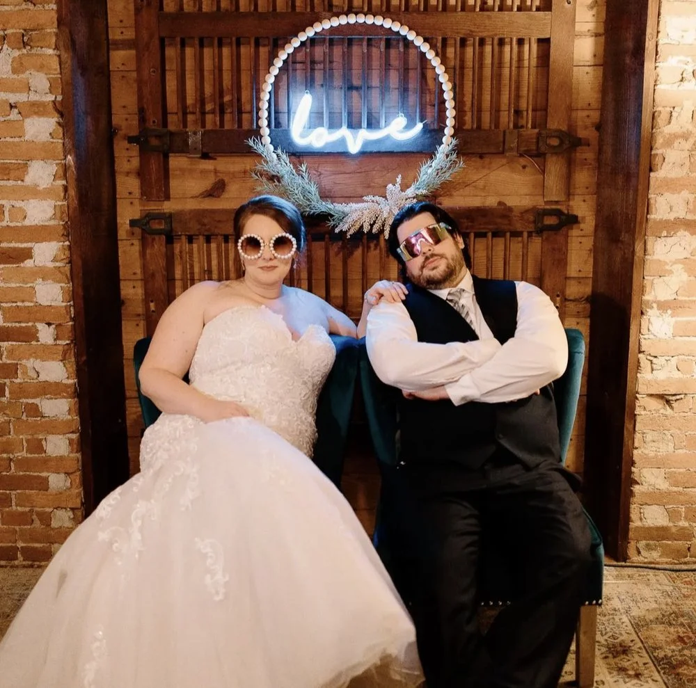 Caitlin and Ben sit side by side on teal velvet chairs beneath a glowing blue neon “love” sign, wearing playful sunglasses at their wedding.