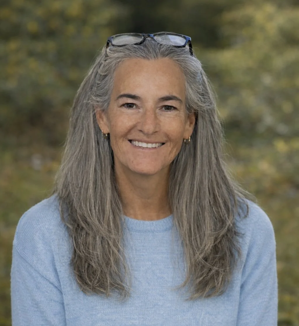 A woman with long brown hair smiling outdoors, wearing a green top and hoop earrings, with greenery in the background.