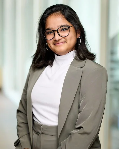 A young woman with glasses, dark hair, and hoop earrings, wearing a gray blazer over a white top, standing indoors with a modern office background.