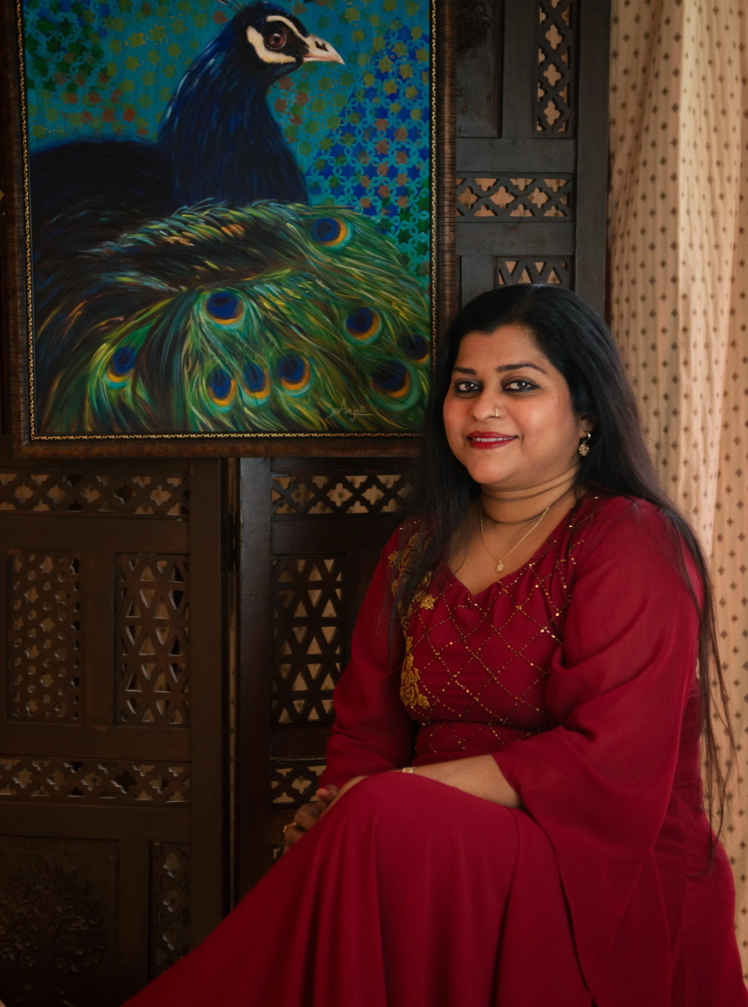 A woman sitting in front of a painting of a peacock with vibrant feathers, wearing a red dress with gold embroidery.