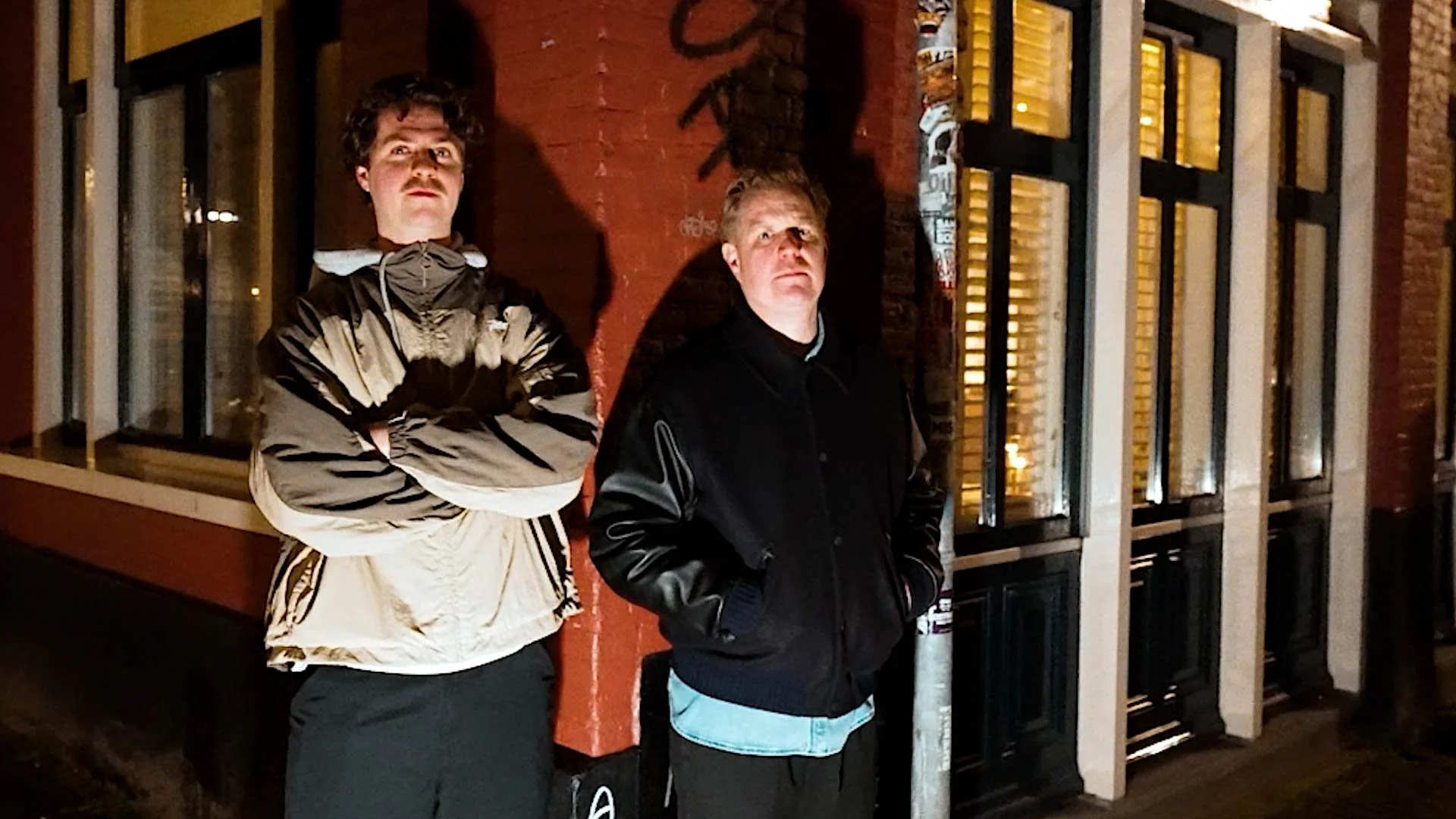 Two young men standing outside at night, one with arms crossed and the other with hands in pockets, in front of a brick wall and large windows with blinds, illuminated by streetlights.