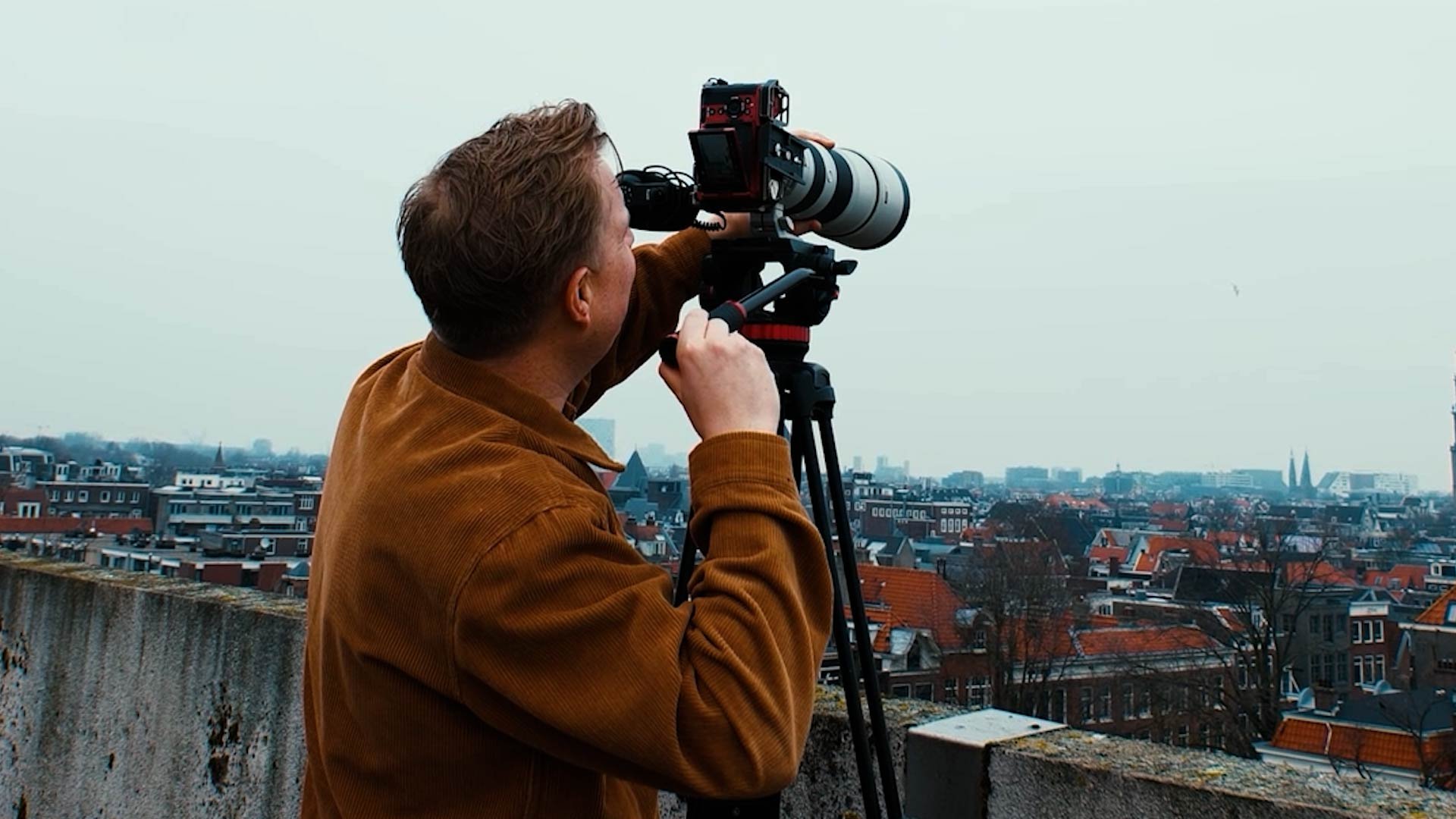 A man in a brown jacket operating a large camera on a tripod on a rooftop, capturing a cityscape of buildings and rooftops under a cloudy sky.