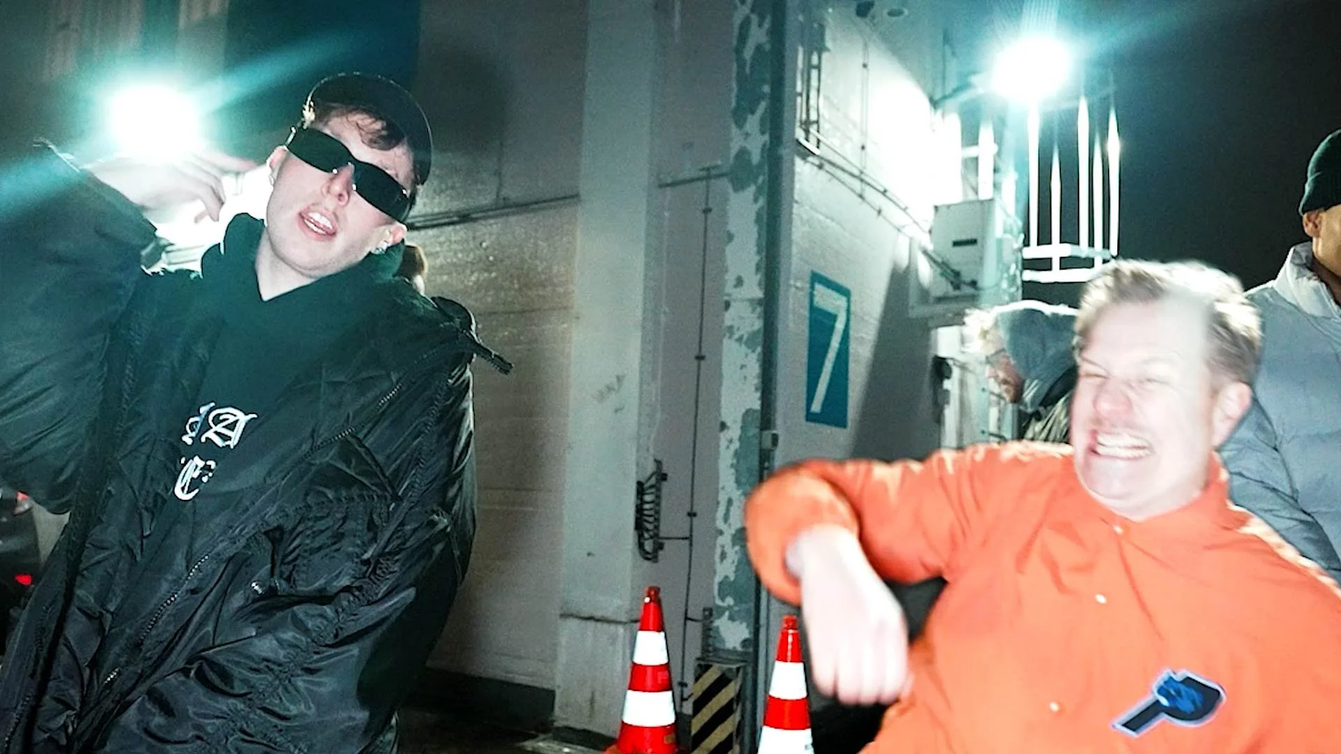 A young man wearing black sunglasses, a black hoodie with white writing, and a black jacket, is making a playful gesture with his hand near his face. Another man in a bright orange shirt is grinning and making a fist gesture. They are outdoors at nig