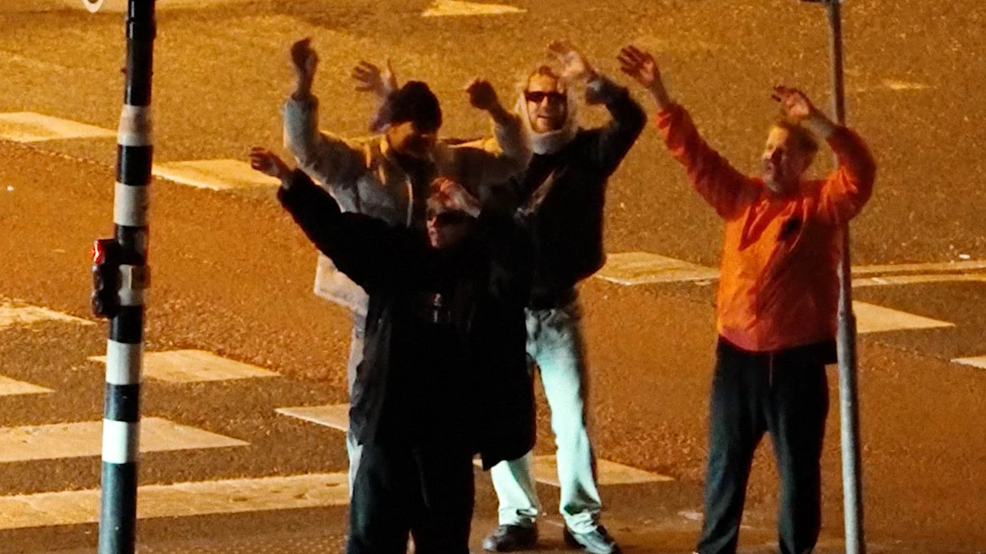 Three young men standing on a street at night, waving their hands, with a poles and pedestrian crossing markings visible in the background.
