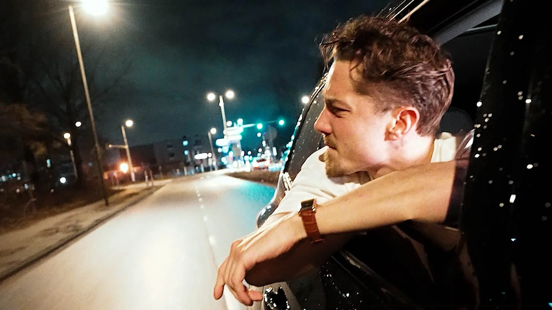 A young man with short brown hair leaning out of a car window during night time, looking relaxed and enjoying the view. The street is illuminated with streetlights and traffic signals in the background.
