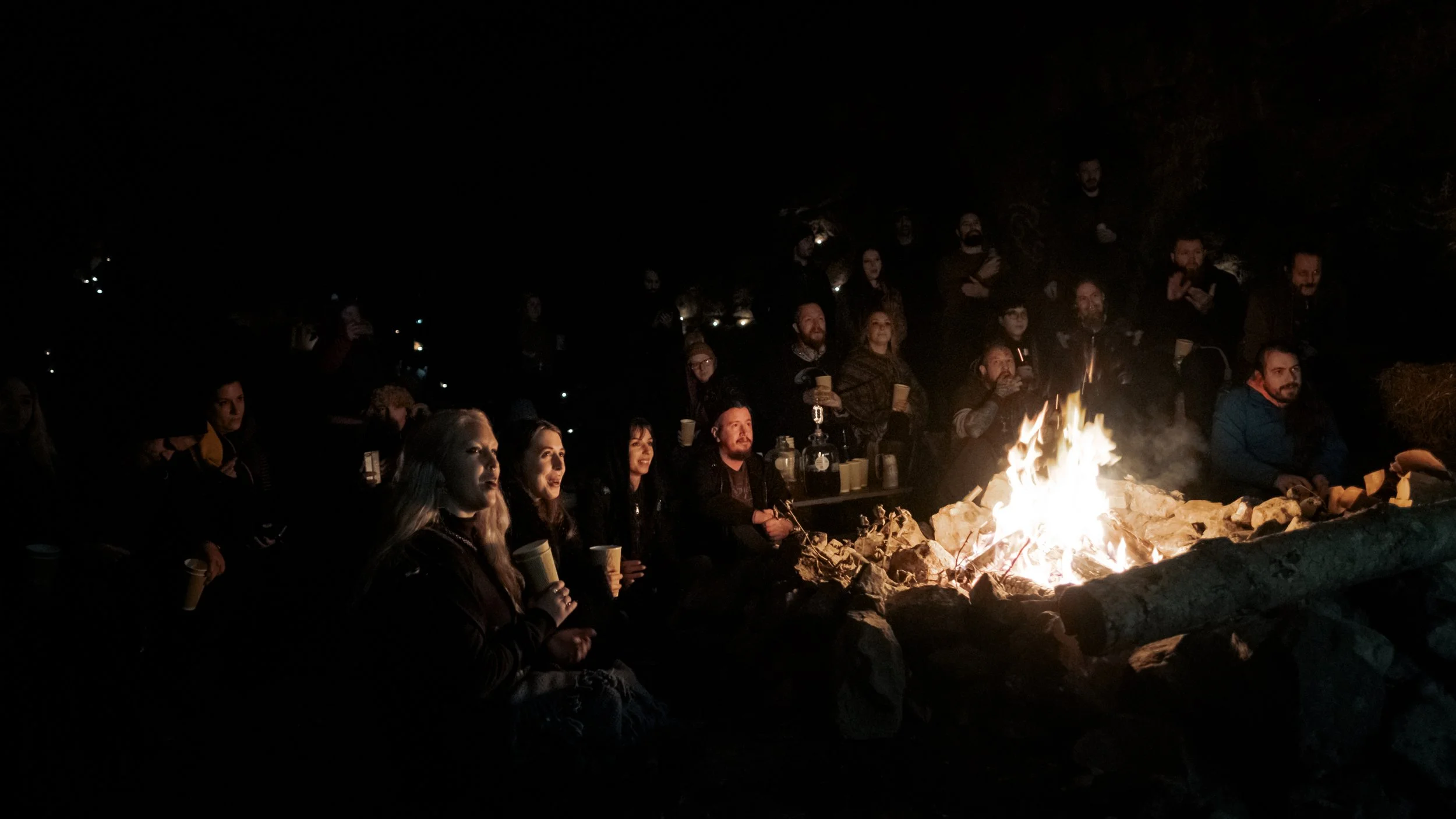A group of people gathered around a fire in a dark cave, which is roaring and seems to captivate those gathered.