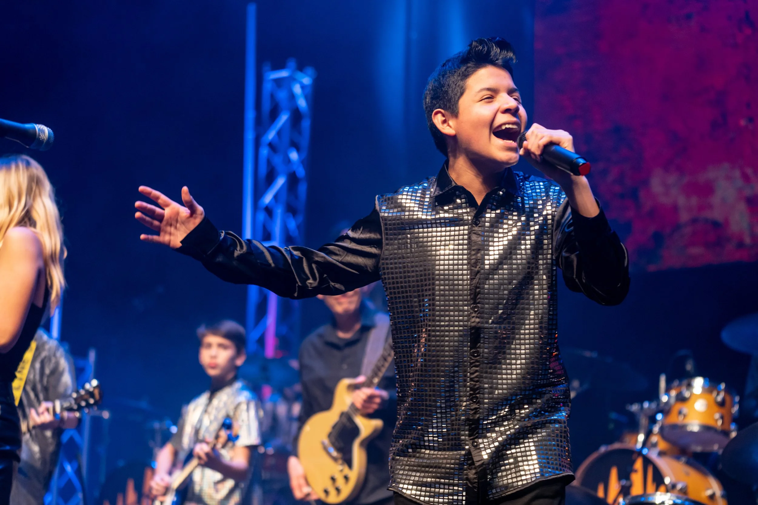 A young vocalist in a silver jacket singing into a microphone on stage, with band members playing behind him under blue stage lights.