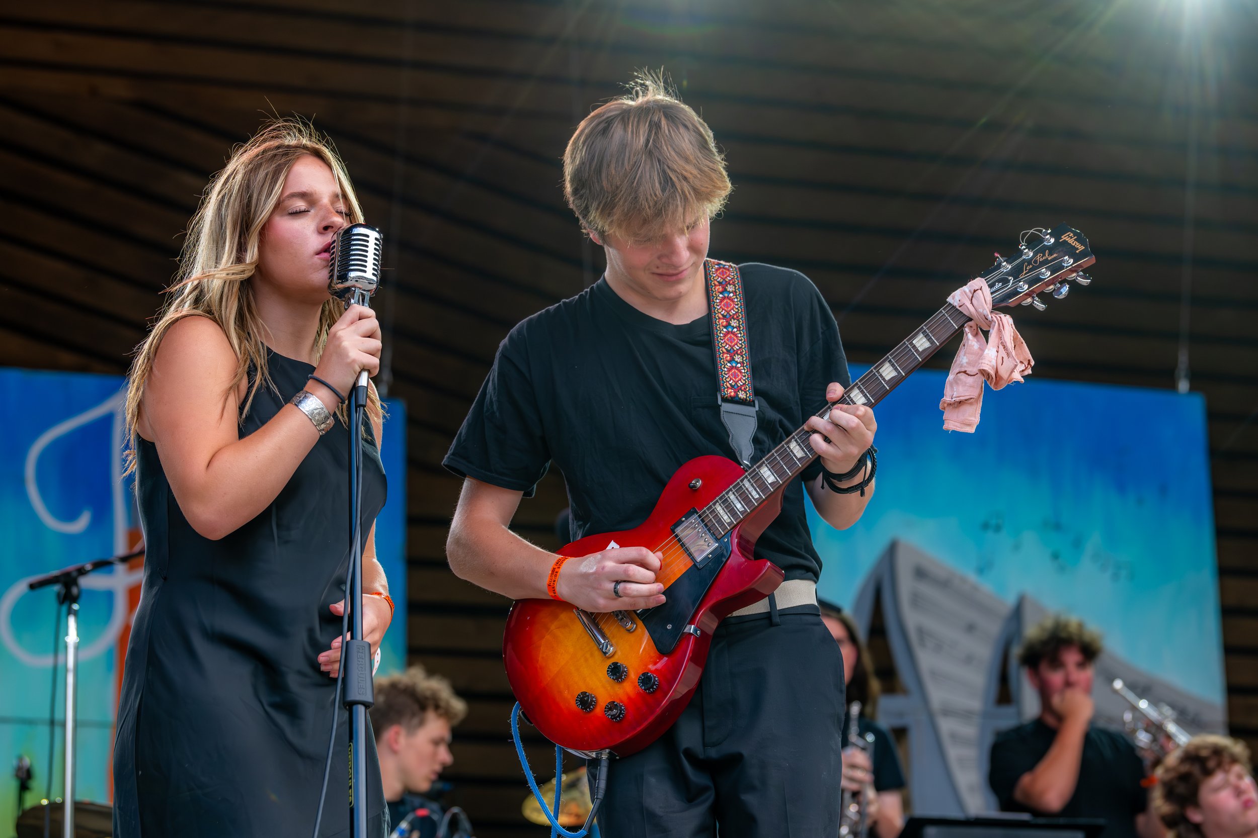 A young woman singing at a microphone beside a teenage guitarist playing a red electric guitar, performing outdoors at a festival.