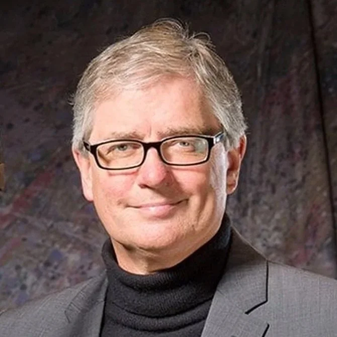Man wearing glasses, a gray suit, and black turtleneck, holding a clarinet, smiling, with a polka dot pocket square.