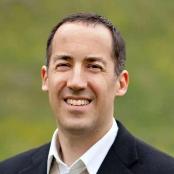 A smiling man with short dark hair, wearing a white collared shirt, beige sweater, and dark blazer, standing outdoors with a blurred green background.