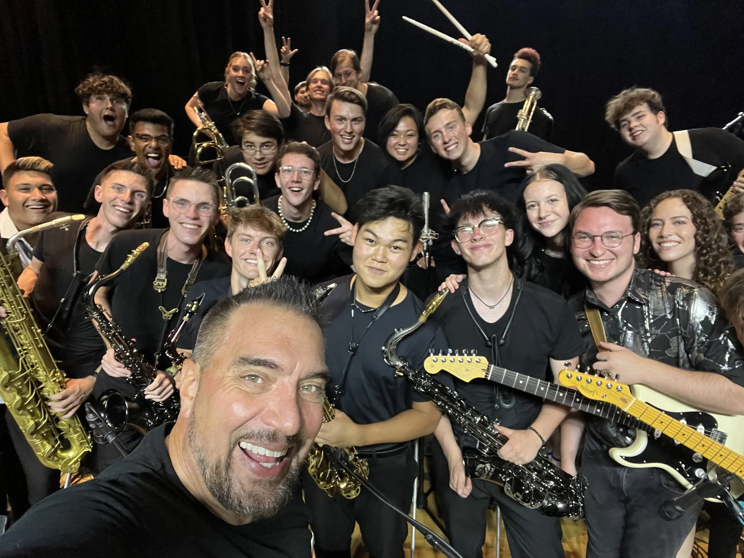 A large group of young musicians crowded together with saxophones, trumpets and guitars, smiling for a group selfie.