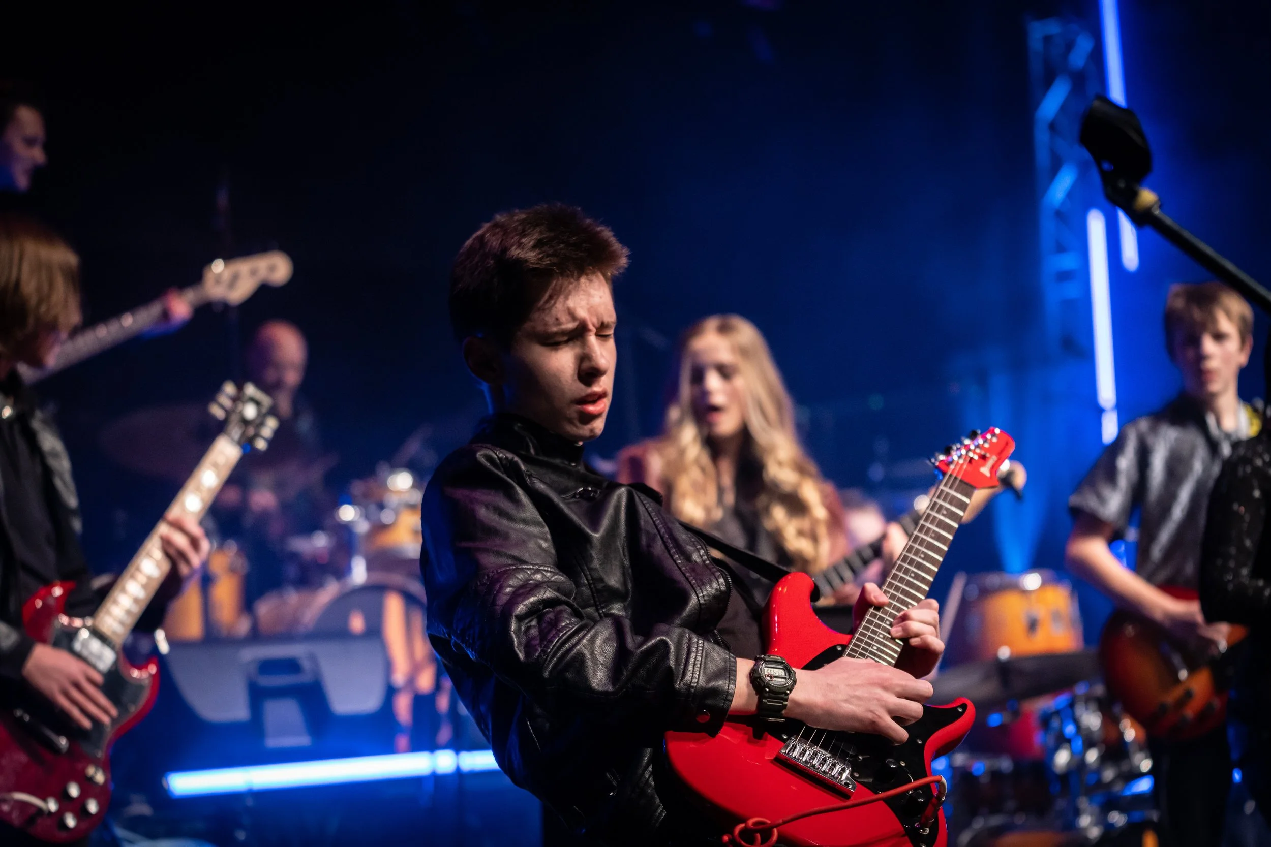 A teenage guitarist playing a red electric guitar on stage with bandmates behind him under blue and purple stage lights.