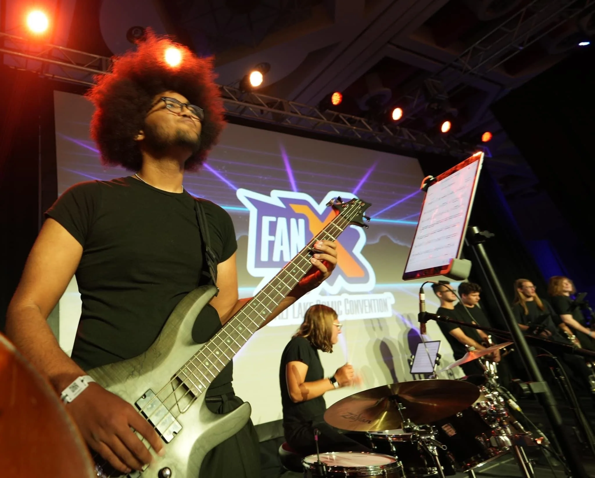 A bassist with curly hair playing an electric bass on stage with a band behind him under colorful stage lights.