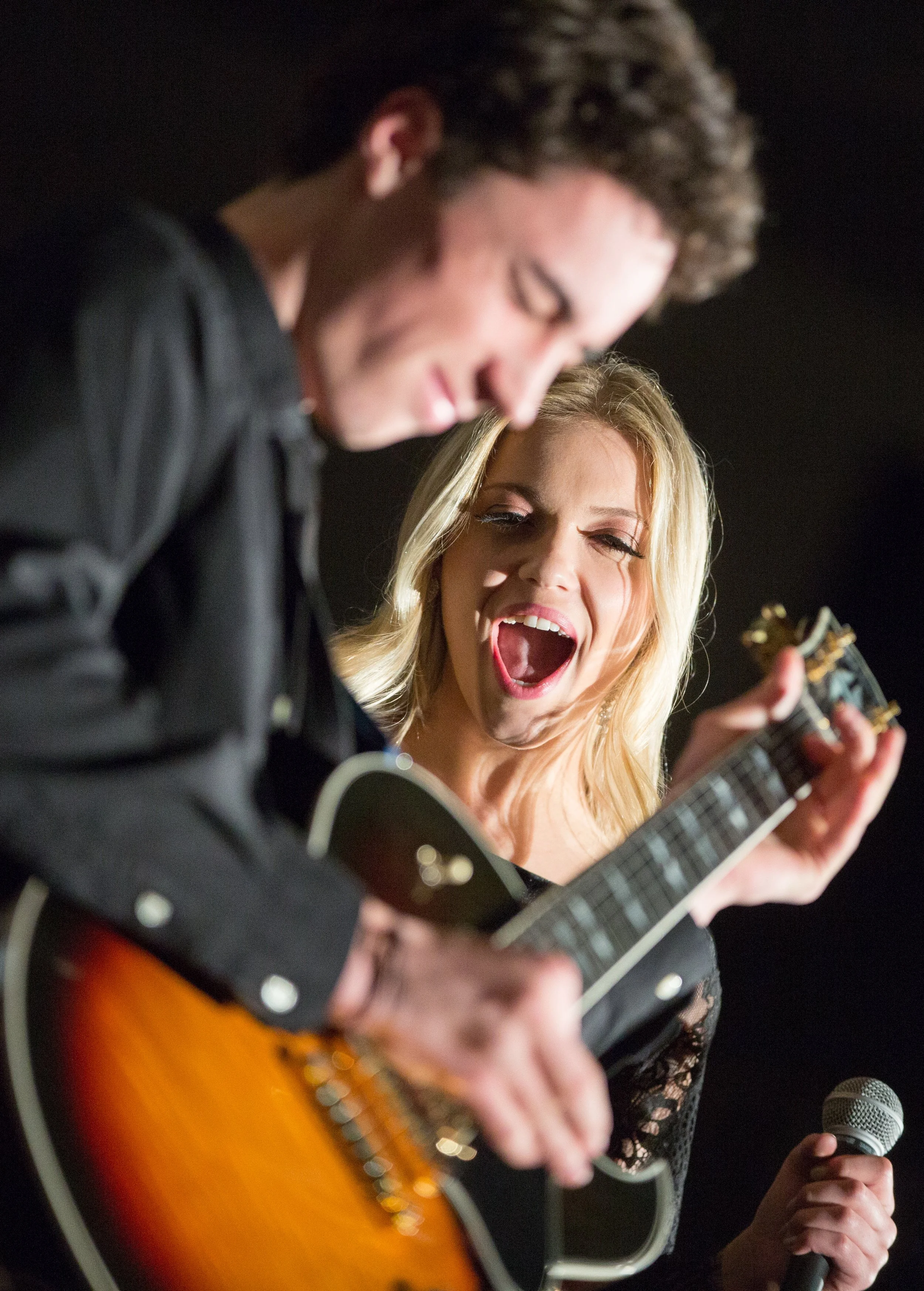 Two young musicians performing together, a guitarist and a vocalist leaning in close under stage lights.