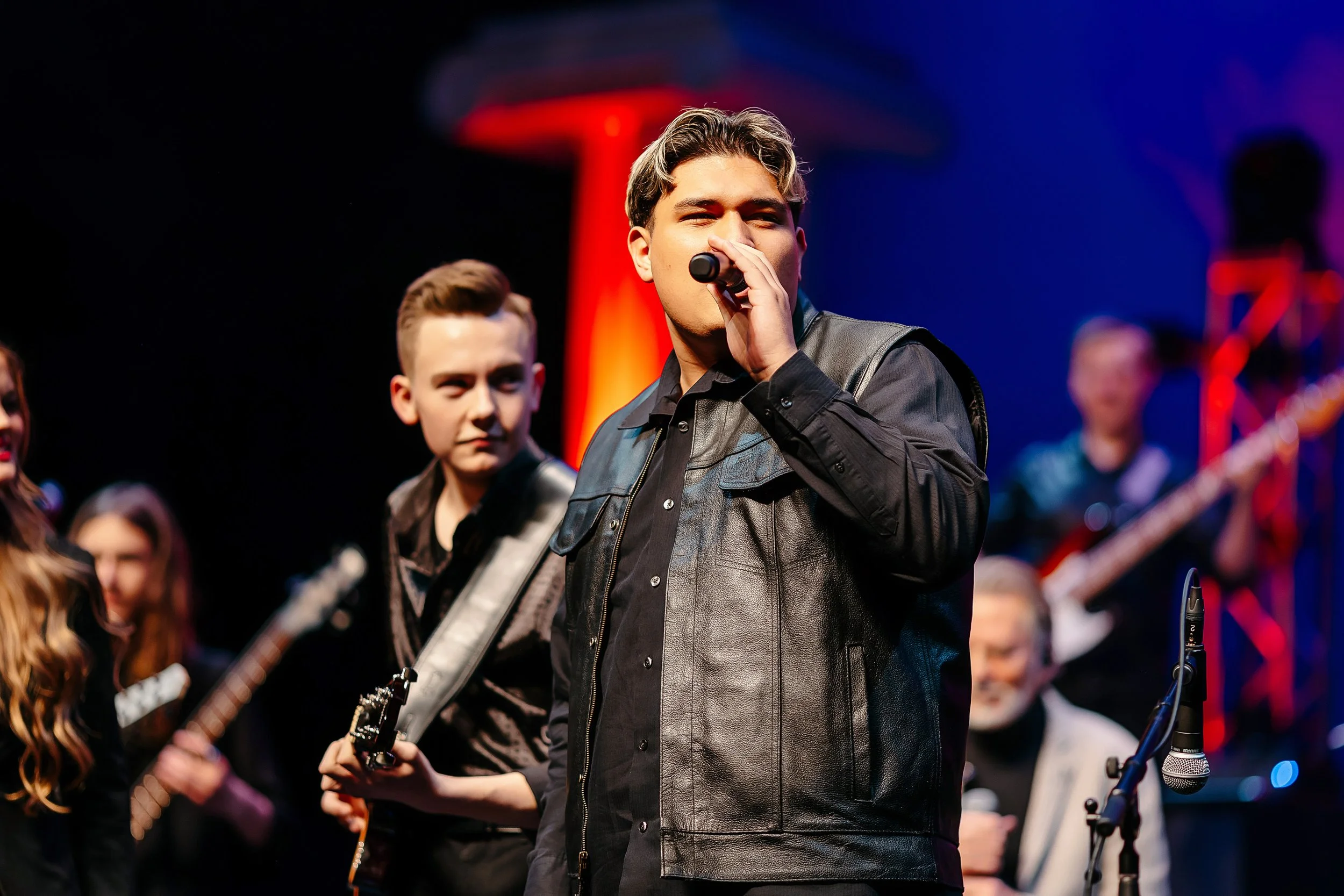 A vocalist singing into a microphone on stage with a band behind him, lit in blue and red stage light.