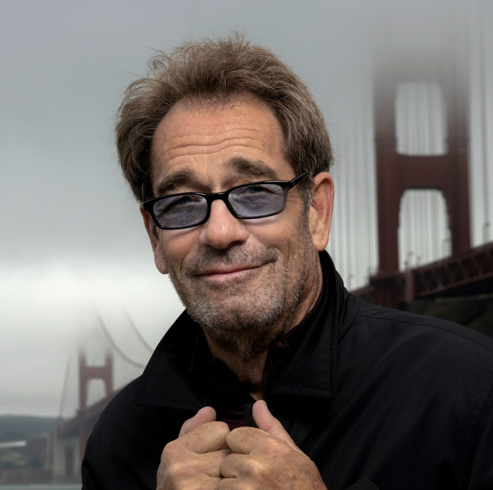 Huey Lewis, a smiling man in sunglasses and a dark jacket, with the Golden Gate Bridge behind him.