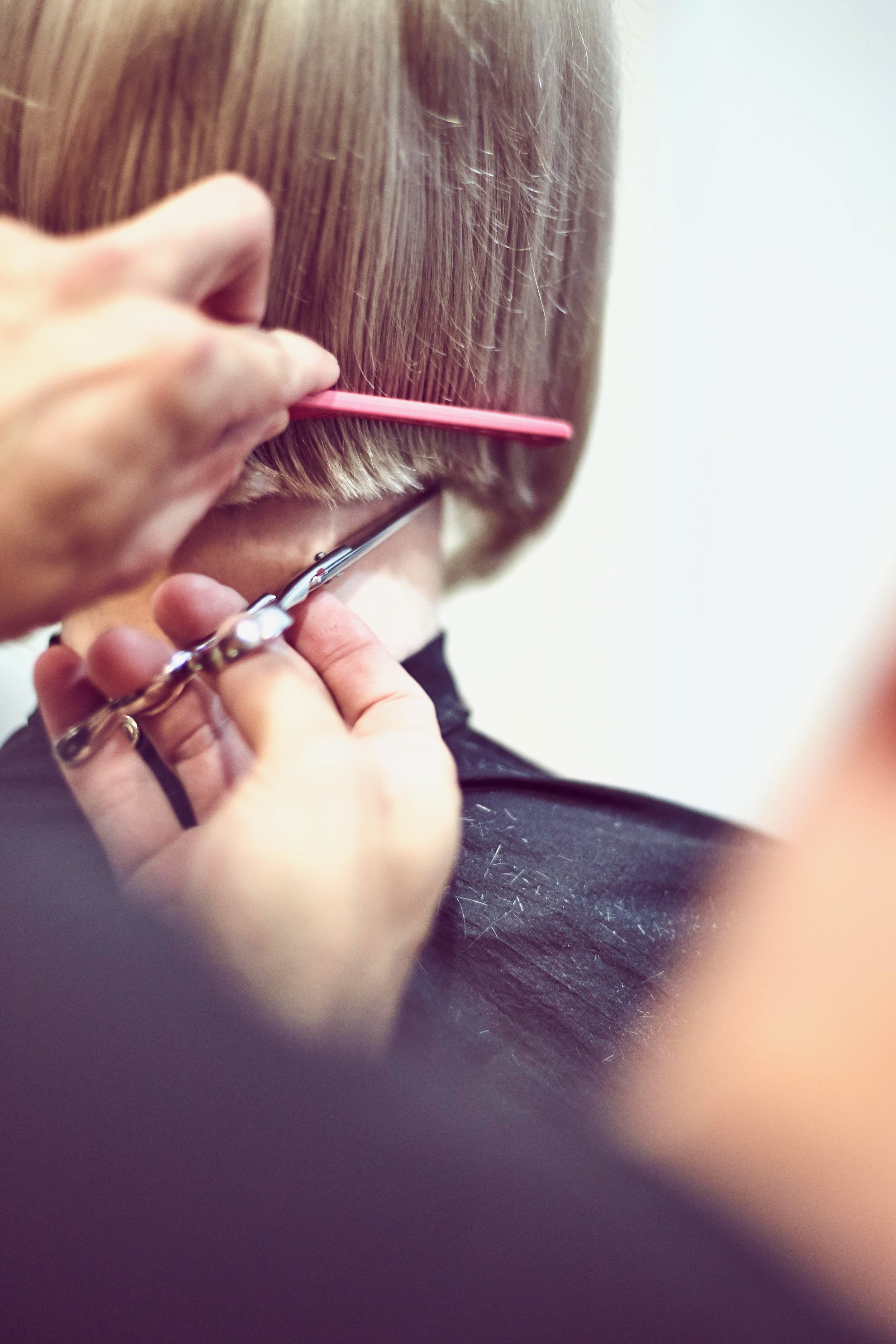 Close-up photo of a person getting a haircut with scissors and a pink comb, focusing on their hair and the barber's hands.
