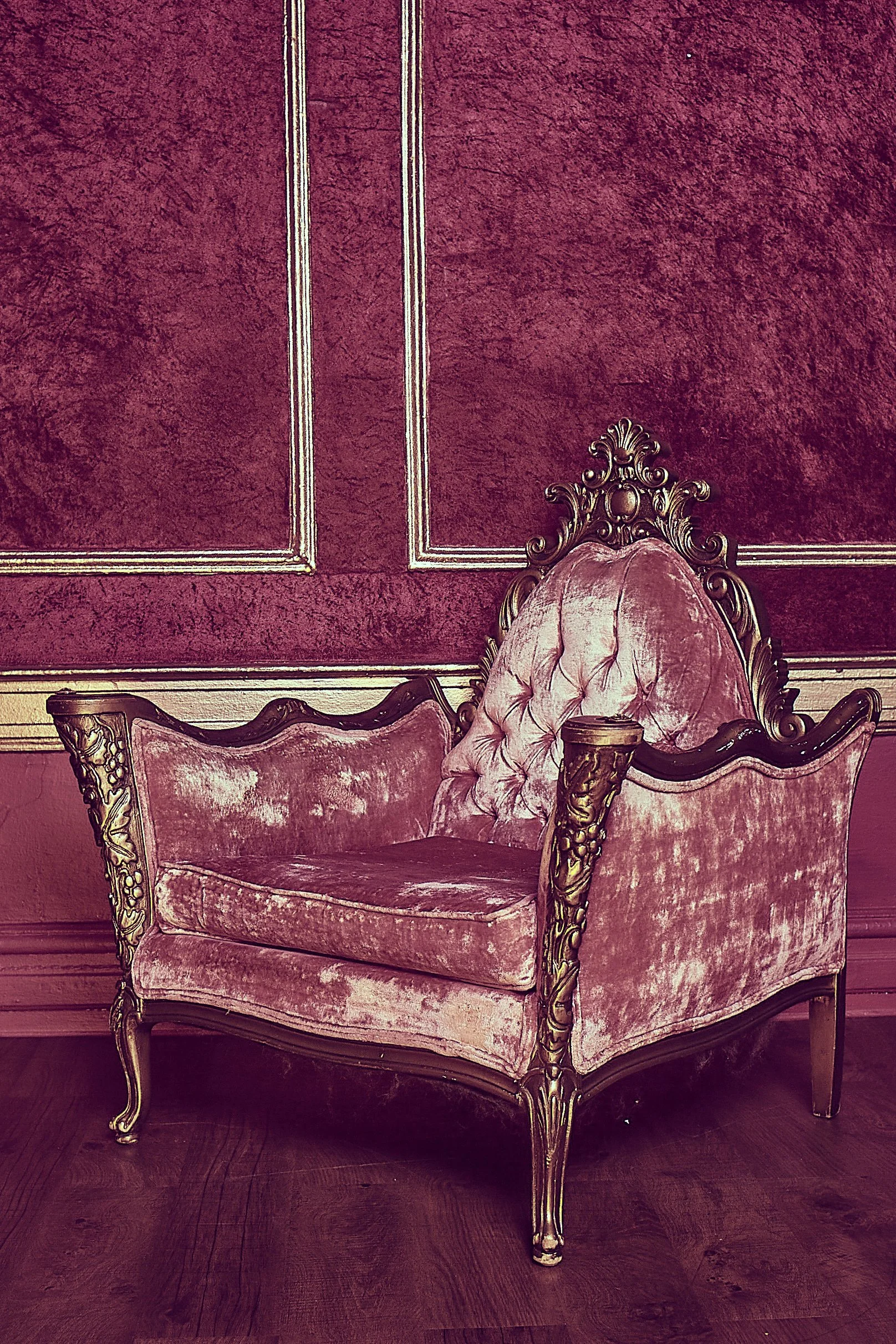 An ornate pink velvet armchair with gold accents on wood frame, set against a matching pink textured wall with decorative trim.